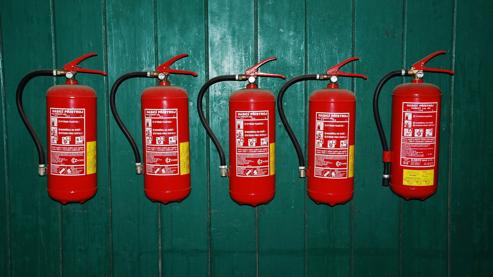 Cinco extintores de incêndio vermelhos alinhados e fixados em uma parede de madeira verde