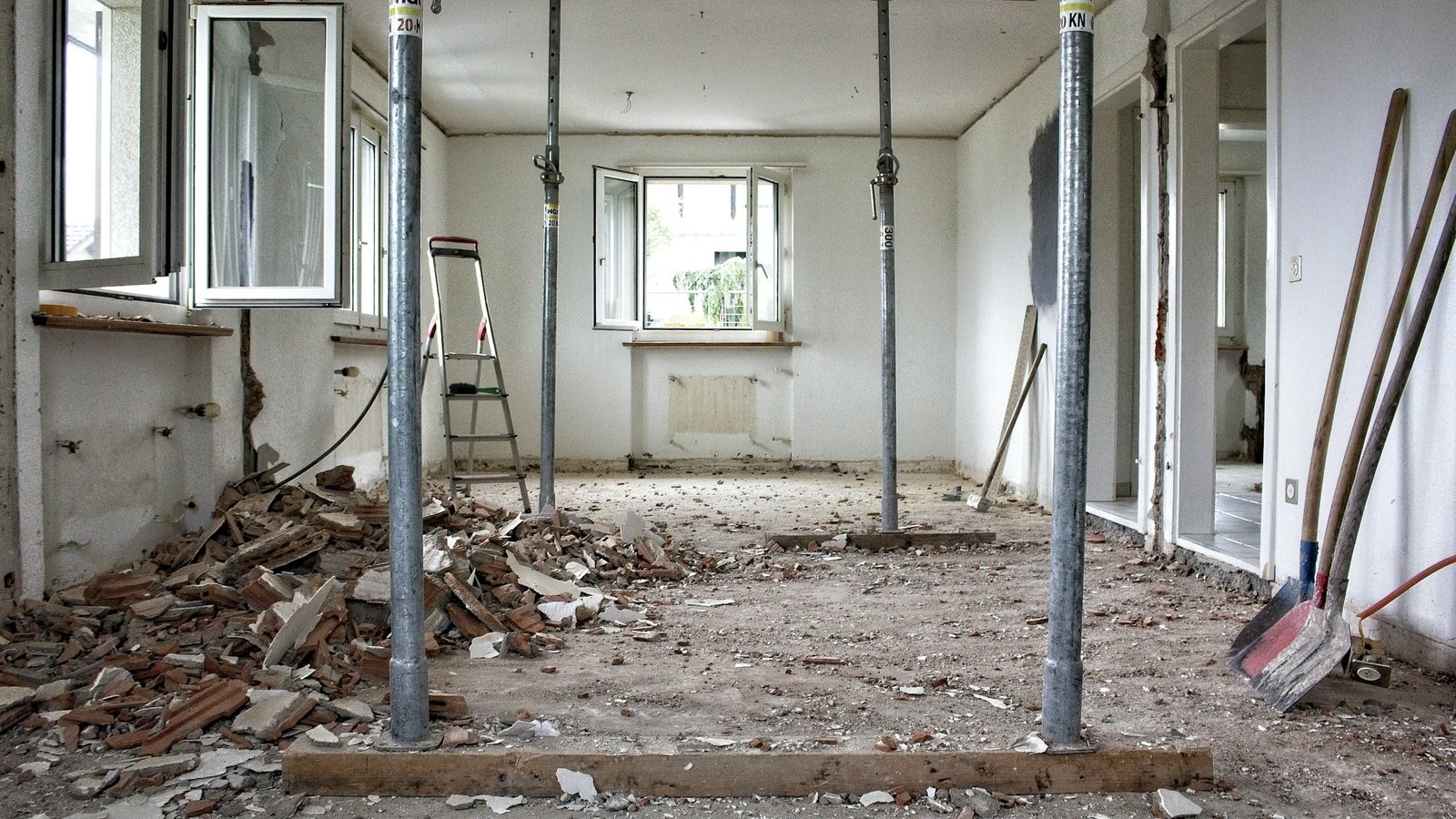 Sala de imóvel em reforma, com escoras metálicas, entulho de alvenaria no piso e ferramentas encostadas na parede