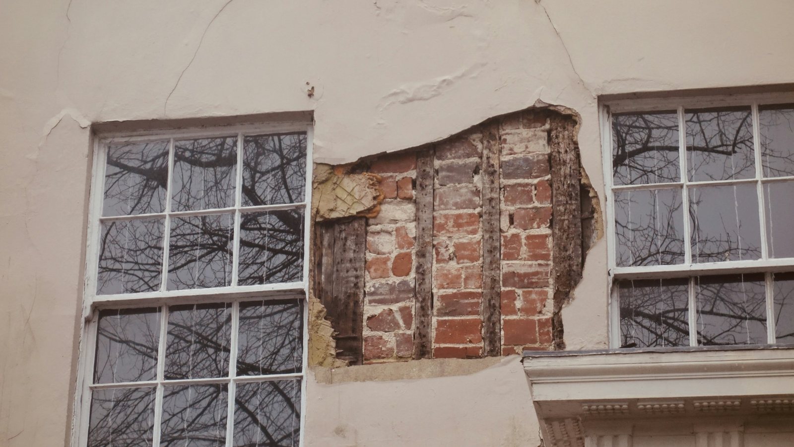 Fachada com fissuras no reboco e trecho de revestimento descolado, que deixa a alvenaria exposta entre duas janelas
