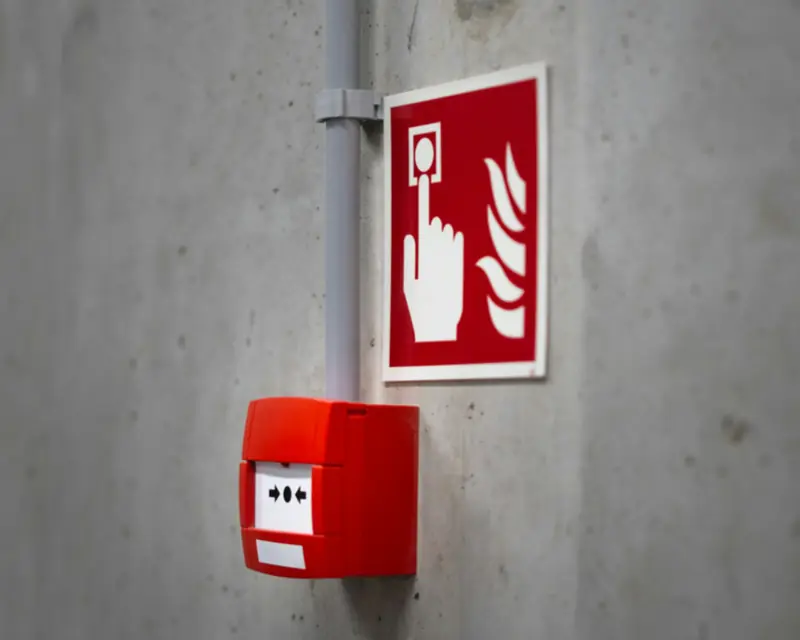Acionador manual de alarme de incêndio, vermelho, em parede de concreto, com placa de sinalização acima