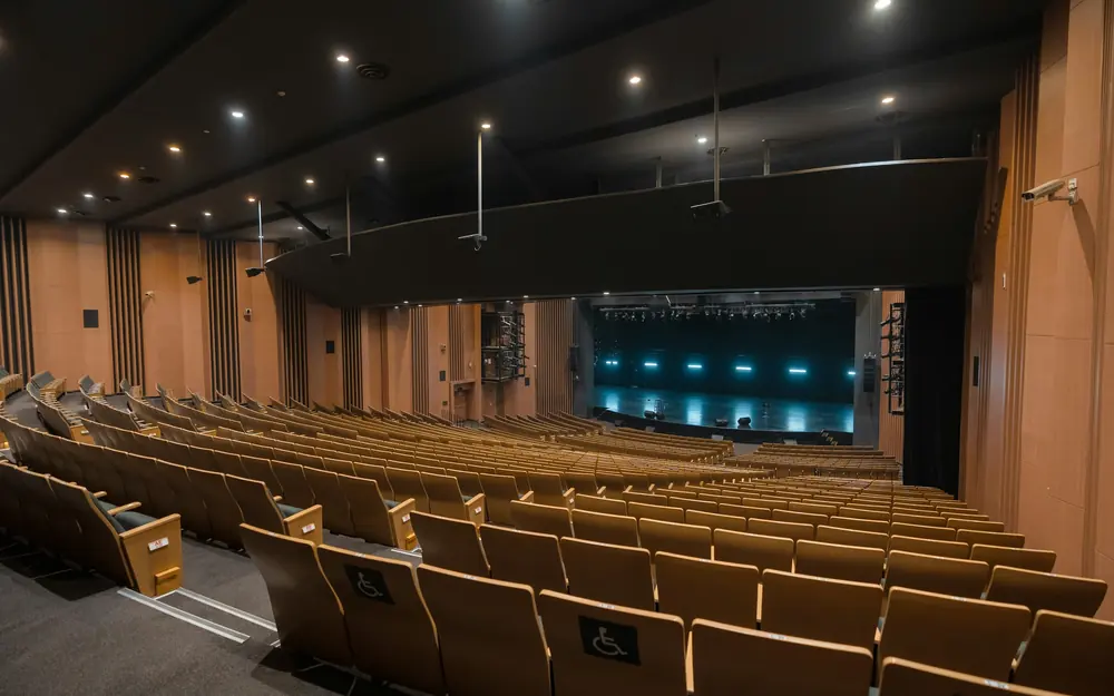 Auditório vazio com fileiras de poltronas de madeira voltadas para o palco iluminado
