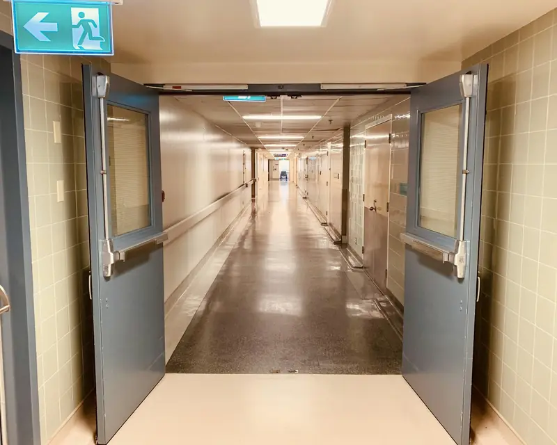 Corredor com porta dupla aberta, barras antipânico nas folhas e placa de saída com seta e figura correndo