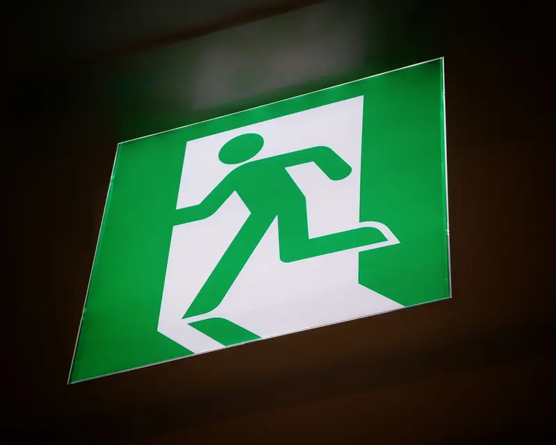 Placa iluminada de saída de emergência, verde, com figura correndo em direção a uma porta