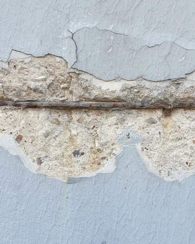 Trecho de parede de concreto pintada com o cobrimento desplacado e a barra de aço aparente e oxidada